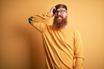 Wall Mural - Handsome Irish redhead man with beard wearing glasses over yellow isolated background smiling confident touching hair with hand up gesture, posing attractive and fashionable