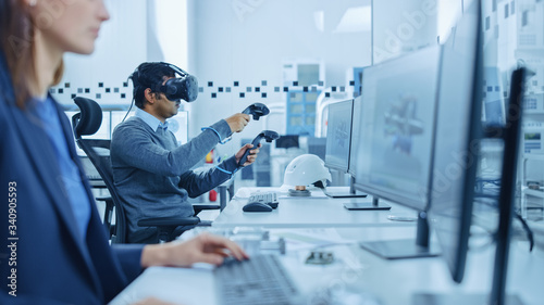 Modern Industrial Factory: Mechanical Engineer Wearing Virtual Reality Headset, Holding Controllers, Uses VR technology for Industrial Design, Development and Prototyping in CAD Software on Computer.