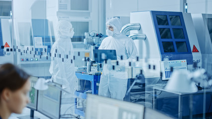 In Clean Workshop: Professional Workers in Coveralls and Masks Use Laptop Walk, Talk. Inside Factory Office: Female Electronics Engineer Works on Computer, Screen Shows Printed Circuit Board 3D Design