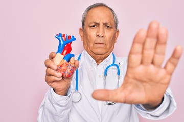 Sticker - Senior hoary doctor man wearing stethoscope holding plastic heart over pink background with open hand doing stop sign with serious and confident expression, defense gesture