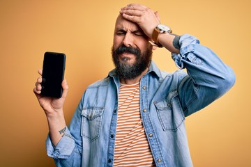 Sticker - Handsome bald man with beard holding smartphone showing screen over yellow background stressed with hand on head, shocked with shame and surprise face, angry and frustrated. Fear and upset