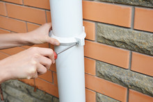 Rain Gutter Installation: A Man Is Screwing The Downpipe Bracket, Socket Clip With A Screwdriver To Fix The Downspout To The Wall Of A Brick House.