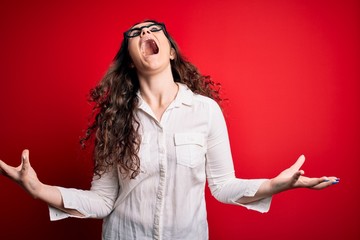 Poster - Young beautiful woman with curly hair wearing shirt and glasses over red background crazy and mad shouting and yelling with aggressive expression and arms raised. Frustration concept.