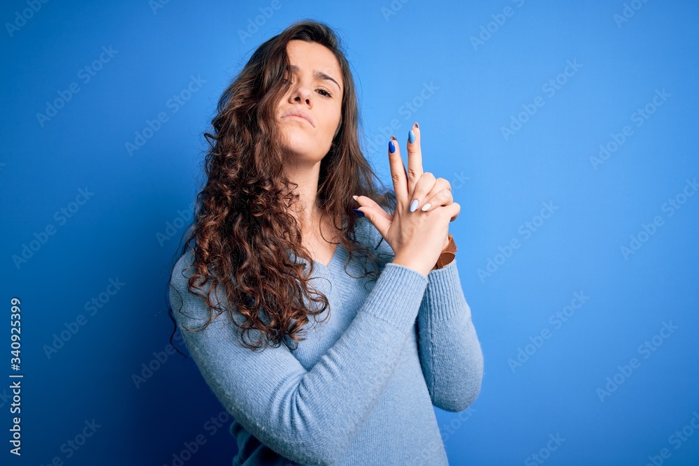 Young beautiful woman with curly hair wearing blue casual sweater over isolated background Holding symbolic gun with hand gesture, playing killing shooting weapons, angry face