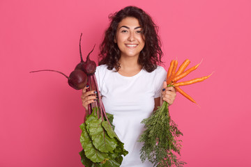 Close up portrait of hungry pleasant sweet brunette standing isolated over pink background, holding raw vegetables, having beet and carrots for meal, wearing white t shirt. People and vegetarianism.