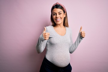 Sticker - Young beautiful brunette woman pregnant expecting baby over isolated pink background success sign doing positive gesture with hand, thumbs up smiling and happy. Cheerful expression and winner gesture.