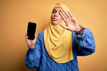 Poster - Young beautiful brunette arab woman wearing islamic hijab holding smartphone showing screen with open hand doing stop sign with serious and confident expression, defense gesture