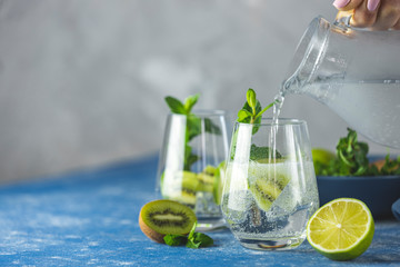 Wall Mural - Women hand is pouring detox water, mojito or martini tonic cocktail from glass jar to glass with kiwi, lime, ice and mint. Summer fresh lime soda cocktail, selective focus.