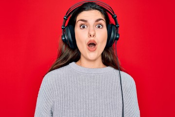 Poster - Young beautiful girl wearing modern headphones listening to music over red background afraid and shocked with surprise expression, fear and excited face.