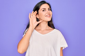 Poster - Young beautiful brunette woman wearing casual white t-shirt over purple background smiling with hand over ear listening an hearing to rumor or gossip. Deafness concept.