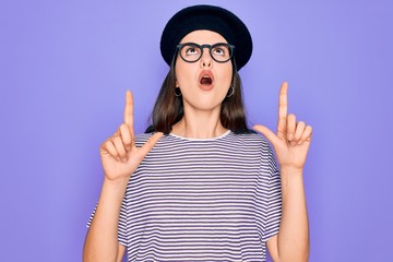 Poster - Young beautiful brunette woman wearing glasses and french beret over purple background amazed and surprised looking up and pointing with fingers and raised arms.