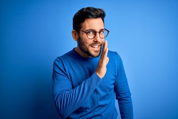 young handsome man with beard wearing casual sweater and glasses over blue background hand on mouth 