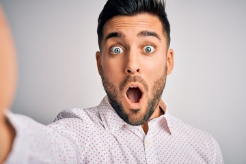 Young handsome man wearing shirt making selfie by the camera over white background scared in shock with a surprise face, afraid and excited with fear expression