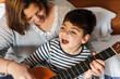 niño juega y aprende con  una mujer a tocar la guitarra en la cama. son madre e hijo, él tiene una pluridiscapacidad. Actividad sensorial. están  alegres y riendo. instrumento de música. educación.