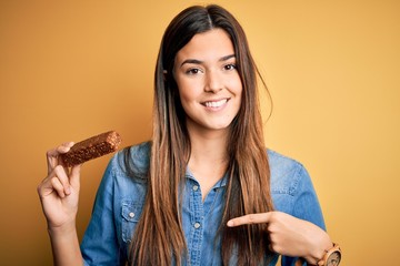 Canvas Print - Young beautiful girl holding healthy protein bar standing over isolated yellow background with surprise face pointing finger to himself