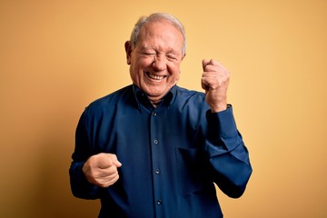 Wall Mural - Grey haired senior man wearing casual blue shirt standing over yellow background celebrating surprised and amazed for success with arms raised and eyes closed. Winner concept.