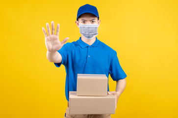 Delivery man wearing mask with carton box picking up the package stop hand gesture to front home receiver shipping deliver cargo prevention infected virus on yellow background isolated studio shot.