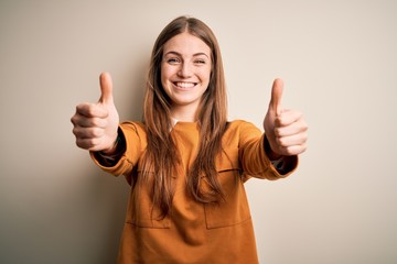 Young beautiful redhead woman wearing casual sweater over isolated white background approving doing positive gesture with hand, thumbs up smiling and happy for success. Winner gesture.