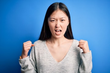 Sticker - Young beautiful asian woman wearing casual sweater standing over blue isolated background angry and mad raising fists frustrated and furious while shouting with anger. Rage and aggressive concept.