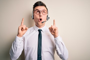 Wall Mural - Young handsome caucasian business man wearing call center headset at customer service amazed and surprised looking up and pointing with fingers and raised arms.