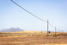 Truck Following Car On Desert Road With Electric Wooden Posts Along In Fuerteventura Island. Arid Landscape With Volcanic Mountain On Background In Canary Islands, Spain