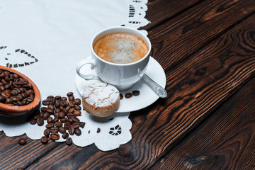 A White cup of hot coffee with cookies on a plate stands on a white napkin on a dark wooden background. Coffee beans are scattered on the table.  A large empty area for advertising text or ideas.