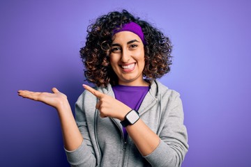 Sticker - Beautiful curly arab sportswoman doing sport wearing sportswear over purple background amazed and smiling to the camera while presenting with hand and pointing with finger.