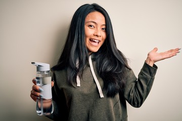 Wall Mural - Young beautiful chinese sportswoman holding bottle of water over isolated white background very happy and excited, winner expression celebrating victory screaming with big smile and raised hands