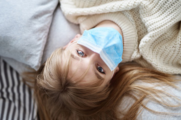 Young attractive teenager blonde girl in white sweater lying on a bed in a medical mask and looking straight to the camera. Coronavirus, quarantine or isolation concept.