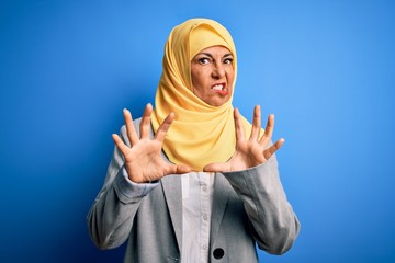 Wall Mural - Middle age brunette business woman wearing muslim traditional hijab over blue background afraid and terrified with fear expression stop gesture with hands, shouting in shock. Panic concept.