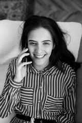 Fashionable young Caucasian woman laying on sofa at home holding mobile phone and talking with friend. She is smiling and enjoying conversation. Black and white photo.