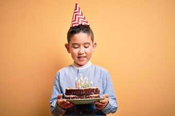 Adorable toddler wearing birthday cap holding cake with candles standing over isolated yellow background