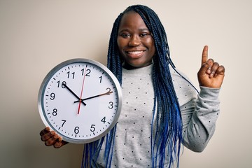 Sticker - Young african american plus size woman with braids doing countdown holding big clock surprised with an idea or question pointing finger with happy face, number one