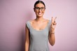 © Krakenimages.com - Young beautiful woman wearing casual striped dress and glasses over pink background smiling with happy face winking at the camera doing victory sign. Number two.