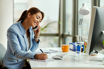 Wall Mural - Happy businesswoman writing notes while talking on the phone in the office.