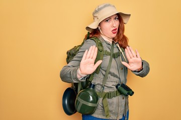 Poster - Young redhead backpacker woman hiking wearing backpack and hat over yellow background Moving away hands palms showing refusal and denial with afraid and disgusting expression. Stop and forbidden.