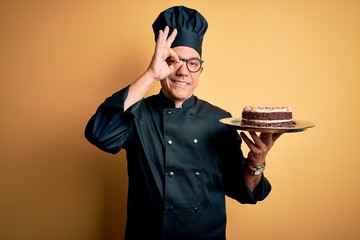 Poster - Middle age handsome grey-haired baker man wearing cooker uniform and hat holding cake with happy face smiling doing ok sign with hand on eye looking through fingers