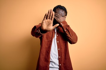 Canvas Print - Young handsome african american man wearing casual jacket standing over yellow background covering eyes with hands and doing stop gesture with sad and fear expression. Embarrassed and negative