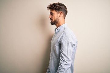 Young handsome man with beard wearing striped shirt standing over white background looking to side, relax profile pose with natural face and confident smile.