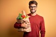 © Krakenimages.com - Young man wearing glasses holding paper bag with food over isolated yellow background with a happy face standing and smiling with a confident smile showing teeth