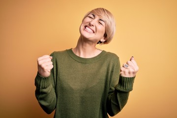 Canvas Print - Young blonde woman with modern short hair wearing casual sweater over yellow background very happy and excited doing winner gesture with arms raised, smiling and screaming for success. Celebration