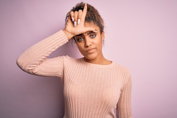 Sticker - Young beautiful african american girl wearing casual sweater standing over pink background making fun of people with fingers on forehead doing loser gesture mocking and insulting.