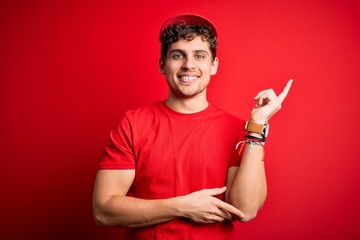 Young blond delivery man with curly hair wearing cap standing over isolated red background with a big smile on face, pointing with hand and finger to the side looking at the camera.