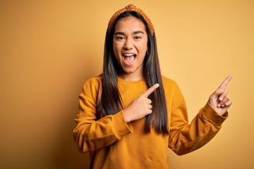 Young beautiful asian woman wearing casual sweater and diadem over yellow background smiling and looking at the camera pointing with two hands and fingers to the side.