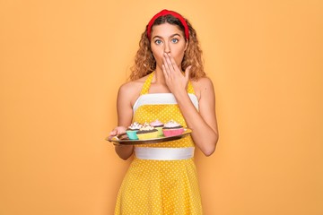 Wall Mural - Beautiful pin-up woman with blue eyes holding chocolate cupcakes over yellow background cover mouth with hand shocked with shame for mistake, expression of fear, scared in silence, secret concept