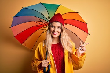 Poster - Young beautiful blonde woman wearing raincoat and wool cap holding colorful umbrella very happy pointing with hand and finger to the side