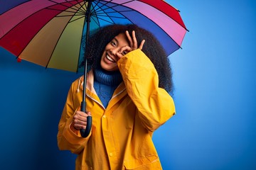 Young african american woman with afro hair under colorful umbrella wearing winter coat for rain with happy face smiling doing ok sign with hand on eye looking through fingers