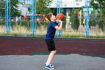 Cute boy in blue t shirt plays basketball on city playground. Active teen enjoying outdoor game with orange ball. Hobby, active lifestyle, sport for kids.