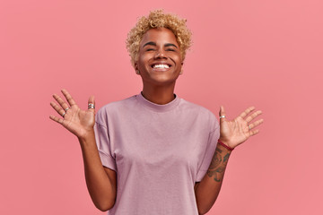 Optimistic black woman raises palms and close eyes from joy, happy to receive awesome present from someone or rejoicing triumph feeling winner, smiles broadly, in a casual lilac t shirt, on pink wall