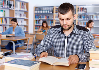 Wall Mural - Portrait of young bearded man sitting in public library and reading books